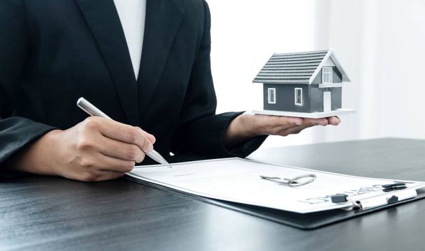 Die Hand des Immobilienmaklers, hält einen Stift in der Hand und entwirft einen Vertrag über den Hausversicherungsvertrag, zusammen mit Mustern von Hausmodellen und Schlüsseln, die er seinen Kunden präsentieren kann.. - Foto, Bild