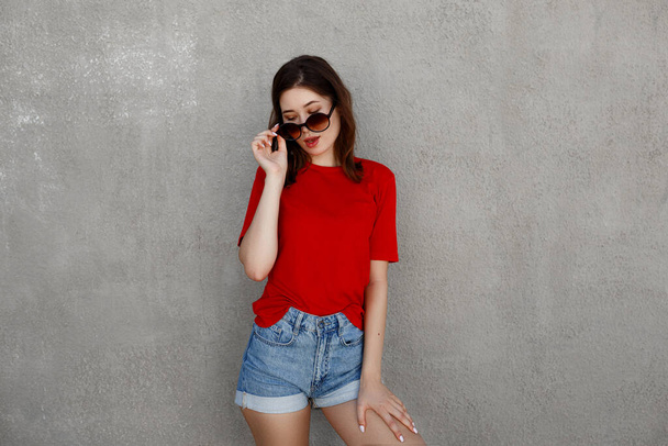 Жіночий азіатський хіпстер у порожній червоній футболці та джинсових шортах, що позують проти грубої вуличної стіни, мінімалістичний стиль міського одягу, макет для друкарні
 - Фото, зображення
