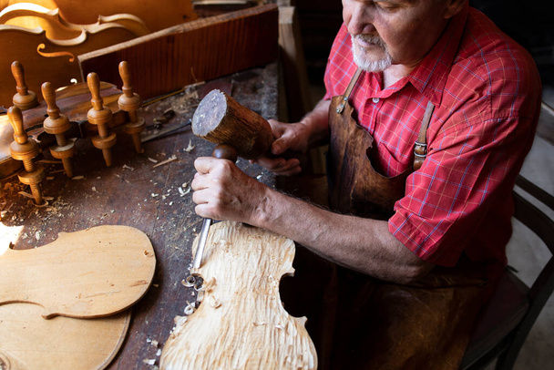 Doświadczony, siwy starszy stolarz pracujący nad swoim projektem w warsztacie stolarskim. Człowiek używający hummera i noża do rzeźbienia deski drewnianej. - Zdjęcie, obraz