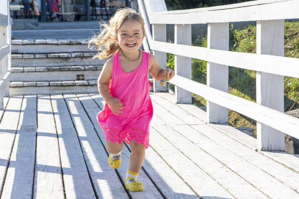 Маленькая блондинка в красном платье бежит по деревянному мосту на фоне городских зданий. - Фото, изображение