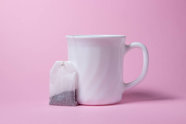 Sac à thé et tasse blanche sur fond rose. Une tasse et un sachet de thé côte à côte. - Photo, image
