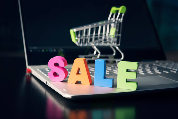 Close up of big colored letters SALE set on opened laptop bottom at shopping cart and black empty display background. Concept of shopping online. Discount in online market place. Laptop screen mockup - Photo, Image