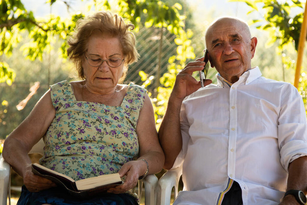 ηλικιωμένο ζευγάρι διαβάζει ένα βιβλίο και μιλάει στο κινητό τηλέφωνο - Φωτογραφία, εικόνα