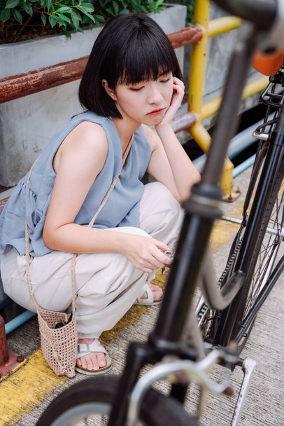 Jonge aziatische Thaise vrouw zitten naast gebroken fiets met deprimerend gevoel wachten tot iemand om het te repareren. - Foto, afbeelding