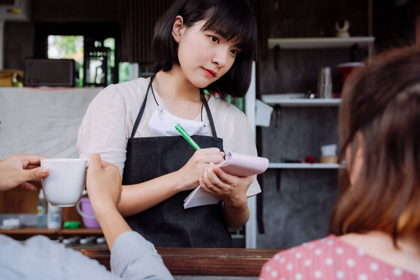 Όμορφη Ασιάτισσα ταϊλανδέζα ιδιοκτήτρια καφετέριας που παίρνει παραγγελία από πελάτη. Τοπικό κατάστημα. Τοπική επιχειρηματίας. - Φωτογραφία, εικόνα
