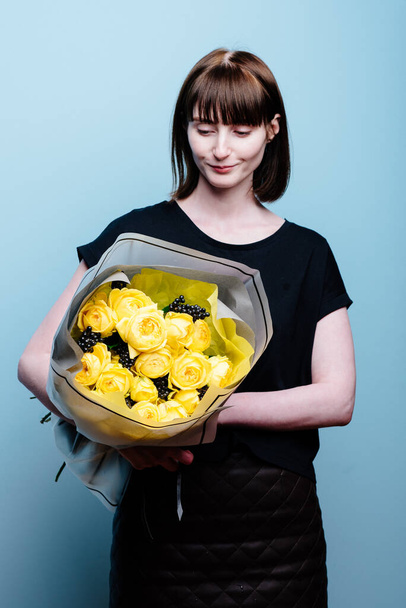 Portrait of young female florist with bouquet of flowers in transparent package in the hands on a blue background . Rosa spray Catalina, rhamnus - Φωτογραφία, εικόνα