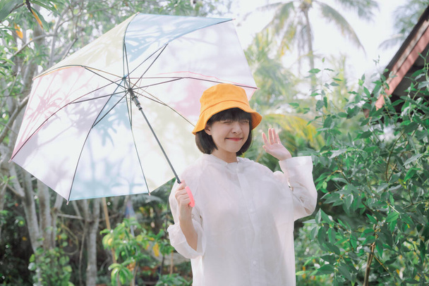 Модний портрет - красива азіатська тайська жінка, коротке темне волосся, одягнена в біле пальто і жовтий капелюх, що тримає парасольку пастельної веселки в сезон дощів
. - Фото, зображення