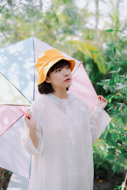 Portret mody - Piękny azjatycki tajski kobieta krótkie ciemne włosy nosić biały płaszcz i żółty kapelusz gospodarstwa pastelowy tęczowy parasol w porze deszczowej. - Zdjęcie, obraz