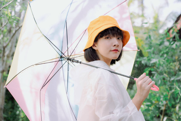 Retrato de moda - Linda mulher tailandesa asiática curto cabelo escuro desgaste casaco branco e chapéu amarelo segurando guarda-chuva pastel arco-íris na estação chuvosa. - Foto, Imagem