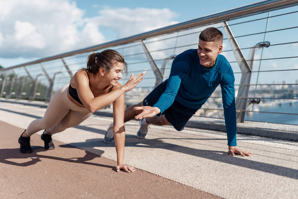 Morning exercise and workout concept. two young happy friends making give five gesture after sport training outdoors, doing push ups together, standing in plank pose - Photo, Image