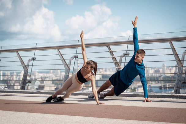 Concept of workout and exercise. Adult sporty woman and man making sport training outdoors, standing in plank pose together, raised hands up - Photo, Image