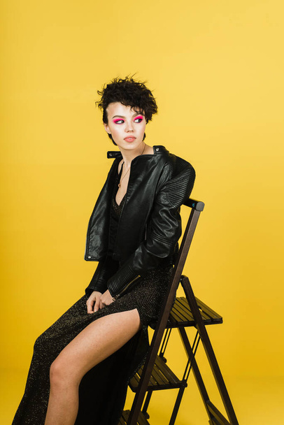 Fashionable portrait of a young girl in a leather jacket and a shiny long dress. Stylish girl in a black rock image on a yellow background in the studio. - Foto, Imagem