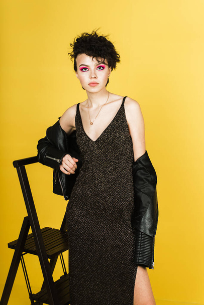 Retrato de moda de una joven con una chaqueta de cuero y un vestido largo brillante. Chica con estilo en una imagen de rock negro sobre un fondo amarillo en el estudio. - Foto, imagen