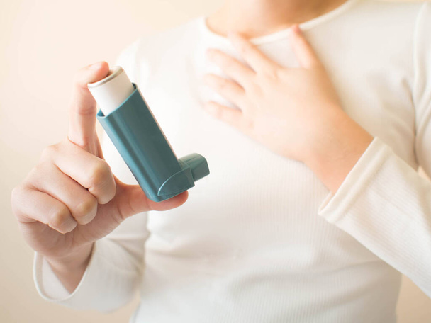 Młoda kobieta w białej koszulce, używająca niebieskiego inhalatora do leczenia astmy. Produkty farmaceutyczne stosuje się w zapobieganiu świszczącemu oddechowi i duszności spowodowanej astmą lub POChP oraz w leczeniu tych objawów. Zamknij się.. - Zdjęcie, obraz