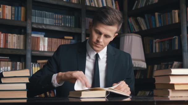 Mladý muž v klasickém obleku sedí v knihovně a hledá informace v knihách - Záběry, video