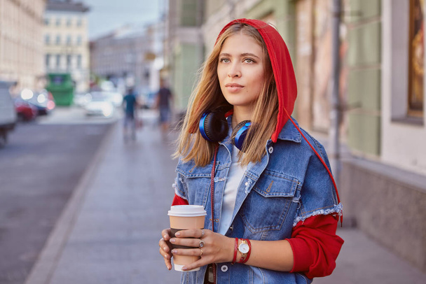 Jovem branca atraente cerca de 25 anos de idade em capuz vermelho e com fones de ouvido está andando no centro de São Petersburgo e bebendo café de copo de papel. - Foto, Imagem