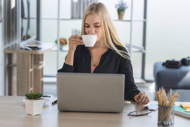Junge Geschäftsfrau, die vom Home-Office aus mit Morgensonne arbeitet. Frau trinkt Kaffee und nutzt Laptop für die Kommunikation in sozialen Netzwerken und die Online-Suche nach Informationen. - Foto, Bild