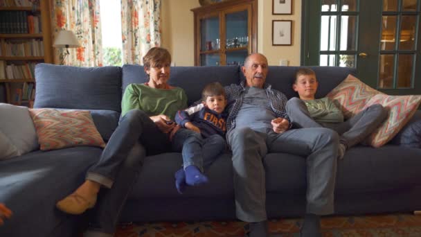 Grandparents and grandchildren sitting on the living room sofa watching television. Home moments in cozy, rustic and old-fashioned house. Full shot. - Footage, Video
