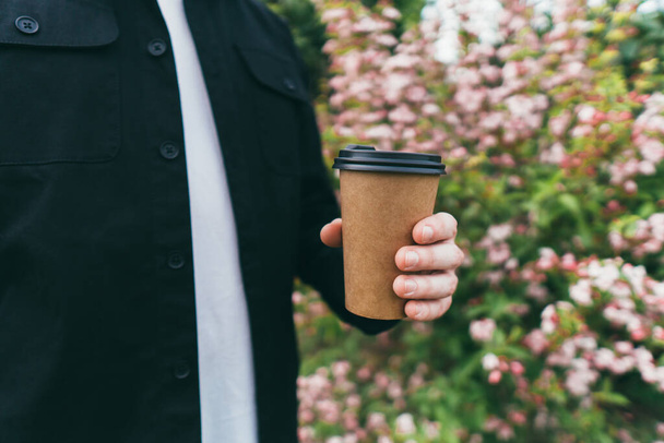 Man holds coffee on the background of beautiful flowers in the park - Fotoğraf, Görsel