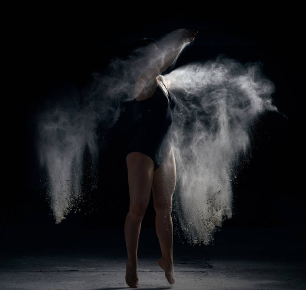 красива біла жінка в чорному боді зі спортивною фігурою танцює в білій хмарі борошна на чорному тлі
 - Фото, зображення