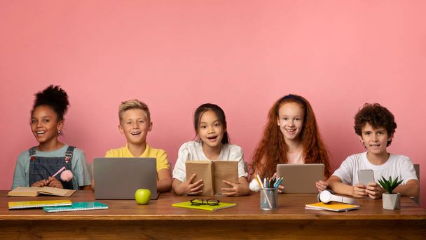 Grupo de alumnos multiétnicos con libros y dispositivos electrónicos sentados a la mesa sobre fondo rosa, espacio en blanco - Foto, imagen