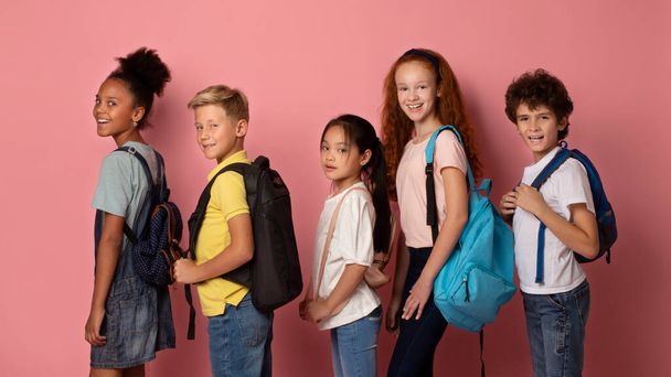 Sırt çantalı neşeli çocuklar pembe arka planda birbiri ardına okula gidiyorlar. - Fotoğraf, Görsel