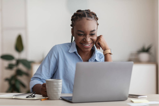 Glimlachende zwarte vrouwelijke werknemer met behulp van laptop en het drinken van koffie op kantoor werkplek - Foto, afbeelding