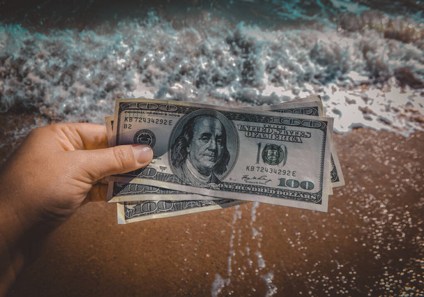 Egy lány, aki 300 dolláros számlát tart a tenger hullámai mögött fehér habbal és homokos strandközeli képpel. Tengerfenéki pénz, tengeri pénz, nyaralás. Koncepció pénzügy pénz nyaralás utazás - Fotó, kép