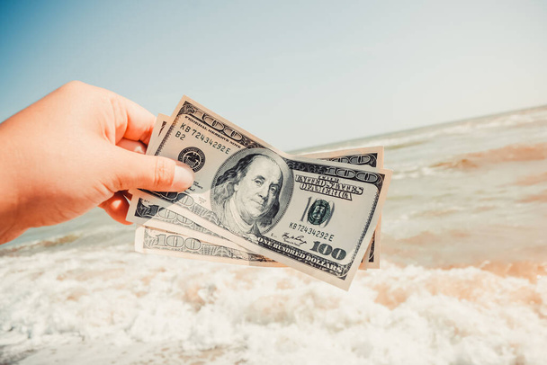 Girl holding a money bill of 300 dollars on background of sea waves and sky on sunny day. Hand waves sea ocean money dollars bills vacation relax summer. Concept finance money holiday horizon skyline - Foto, imagen