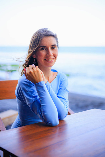 Primer plano retrato de la hermosa mujer caucásica. Mujer sonriente con vestido azul y sentada en el bar de la playa. Estilo de vida. Disfruta la vida. Bali, Indonesia - Foto, imagen