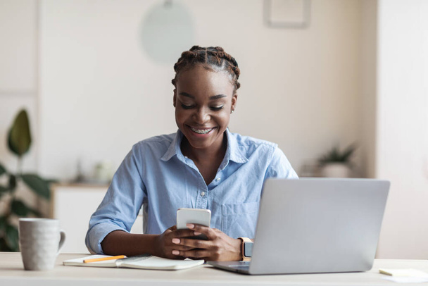 Χαμογελώντας νεαρή αφροαμερικανίδα επιχειρηματίας χρησιμοποιώντας smartphone στο χώρο εργασίας στο γραφείο - Φωτογραφία, εικόνα