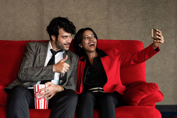 Пара мужчин и женщин фотографируются с билетом, сидя и ожидая перед кинотеатром - Фото, изображение