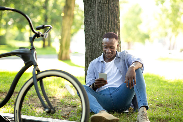 Millennial zwarte man met fiets zitten in de buurt van boom en met behulp van mobiele telefoon in de zomer park - Foto, afbeelding
