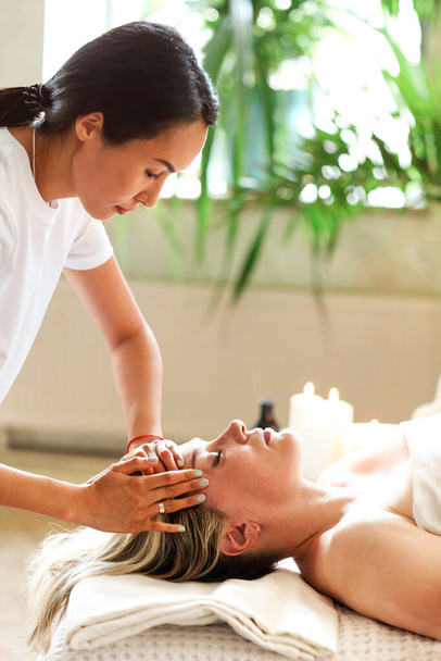 Vista laterale del terapeuta etnico viso massaggiante di donna di mezza età sdraiata sul tavolo da massaggio nel salone spa - Foto, immagini
