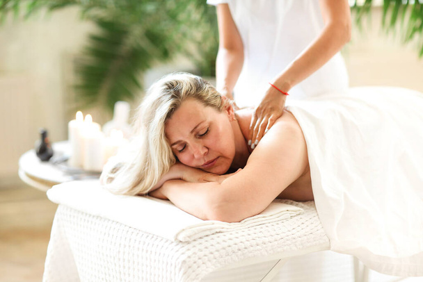 Vue latérale d'une thérapeute ethnique massant le visage d'une femme d'âge moyen allongée sur une table de massage dans un salon de spa - Photo, image