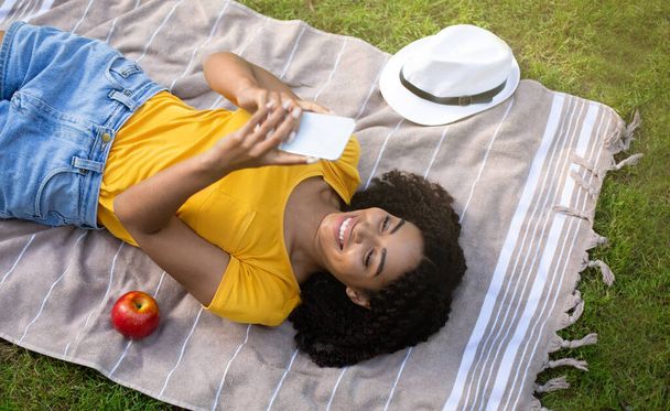 Cool black woman using mobile phone on picnic blanket outside, top view - Photo, Image
