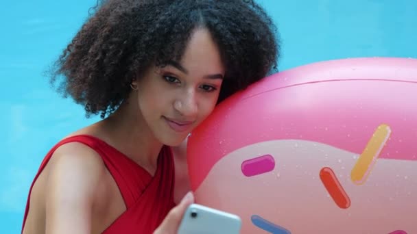 Portrait of playful girl African nationality spending time in pool, emerging from water, hugging inflatable circle in donut form and taking photo with front camera of phone or chatting in video online - Footage, Video