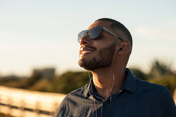Homem latino usando fones de ouvido para ouvir música ou podcasts motivacionais e desfrutar do pôr do sol. Luz quente natural. - Foto, Imagem