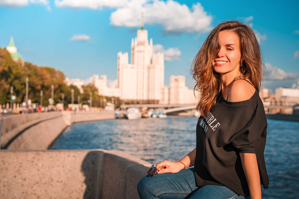 Portrait of a young brunette woman in a jacket with an inscription with the background of the famous high-rise building and the Moscow river on a Sunny summer day - Zdjęcie, obraz