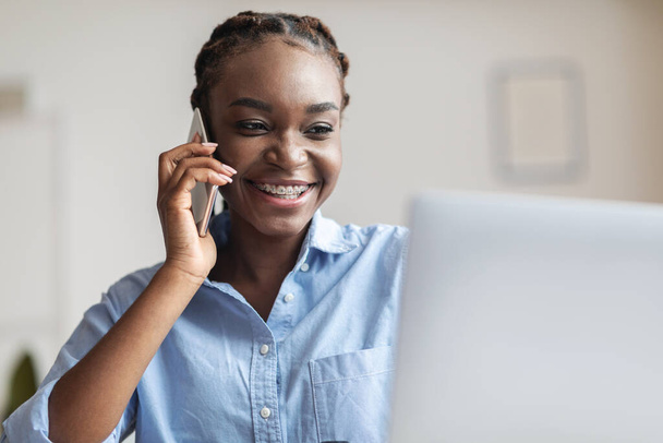 Komunikacja biznesowa. Czarna bizneswoman rozmawiająca przez telefon komórkowy i używająca laptopa w miejscu pracy - Zdjęcie, obraz