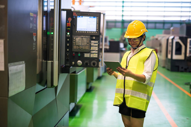 ハードハットと保護メガネを備えたウエストアップ女性労働者は、工場の旋盤ロボットマシンを自動化するためにタブレットの企業アプリをチェックします。検査または監査する技術を有する製造業. - 写真・画像