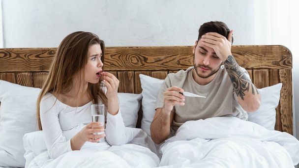 Woman drinking pill, holding glass of water and looking at husband, man touches forehead and looks at thermometer - Photo, Image