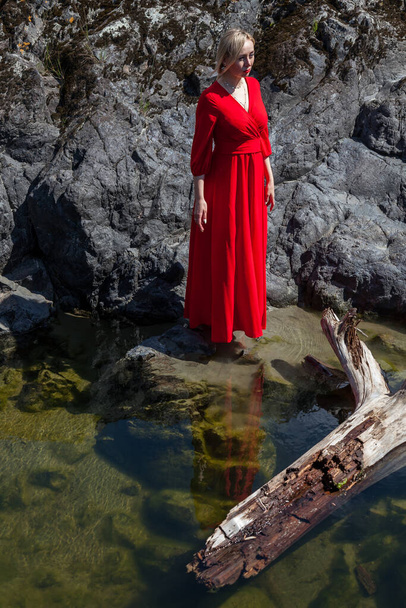 神秘的なイメージの長い赤いドレスのブロンドの女の子はアルタイ山脈の石の海岸の背景に湖の緑の水の上の空気中で急上昇します。魔法と高揚感. - 写真・画像