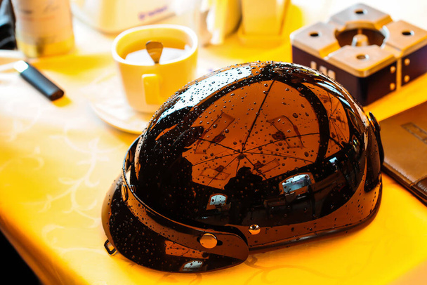 casco de moto negro brillante en estilo vintage con gotas de lluvia puestas en una mesa en un café al lado de una taza de café. - Foto, Imagen