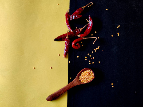 Rode chili vlokken in houten lepel en rode pepers op gele en zwarte achtergrond, bovenaanzicht - Foto, afbeelding