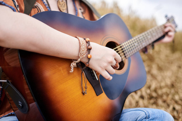 Close-up picture of acoustic guitar held in hands of young hippie women with red hair, wearing boho style clothes, sitting in middle of wheat field,Travel musician in countryside.Musical instrument. - Fotoğraf, Görsel
