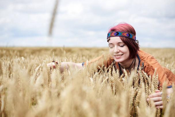 Young woman with red burgundy hair, wearing boho hippie clothes, squatting down in the middle of wheat field. Creative female portrait on natural background. Eco tourism concept. - Foto, Imagen