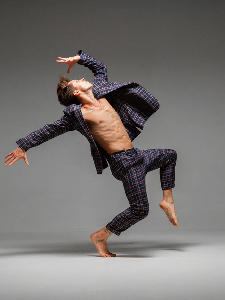 Stylish young guy breakdancer in suit with naked torso and barefoot dancing in studio isolated on gray background - Foto, Bild