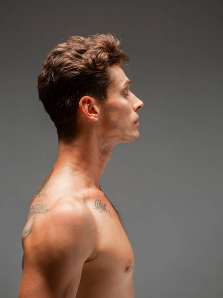 Πορτρέτο του νεαρού άνδρα με κομψό κούρεμα με γυμνούς ώμους που στέκεται στο στούντιο. Σώμα με τατουάζ - Φωτογραφία, εικόνα
