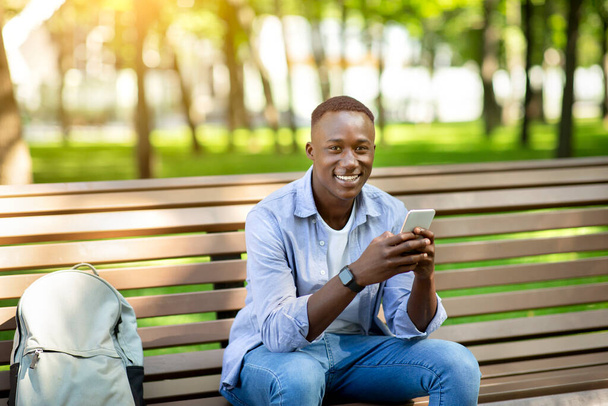 Portrait of African American guy using mobile phone on bench at summer park - Foto, imagen